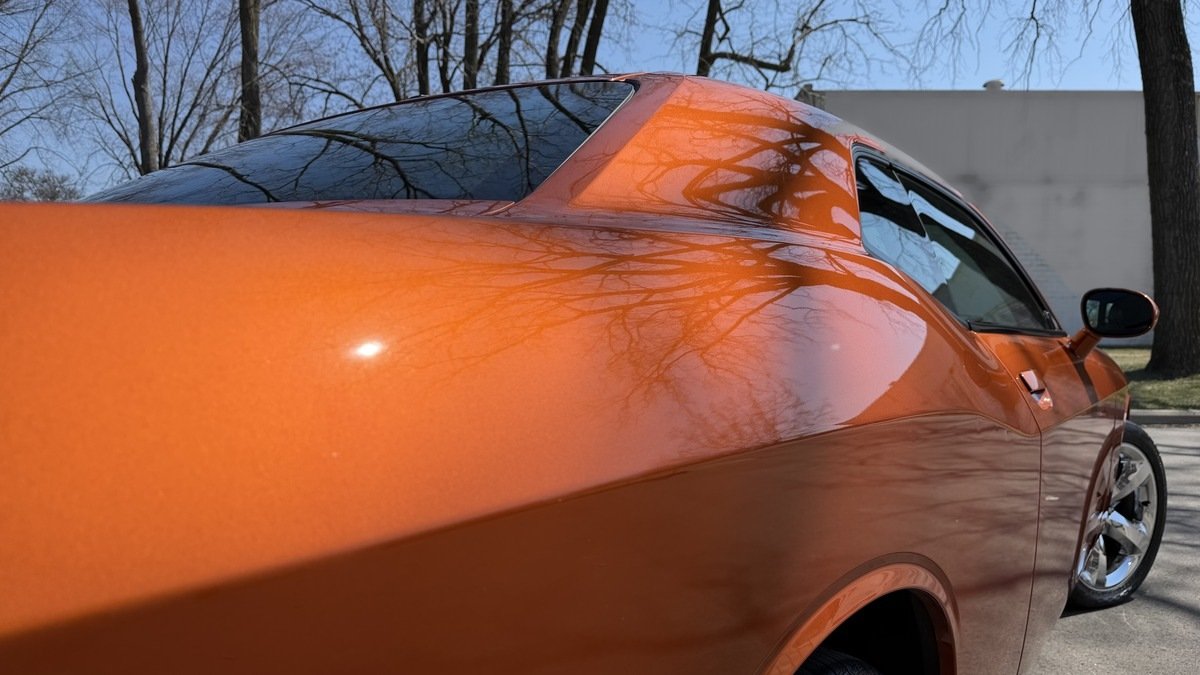 Ceramic Coating Guide 1 – What A Ceramic Coating Actually Does Cover Gloss orange Dodge Challenger with bare trees mirrored in its ceramic coated rear quarter panel.